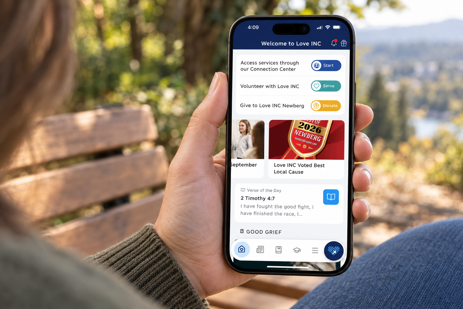Illustrative image of someone holding a phone displaying the Love INC Newberg home screen outdoors.